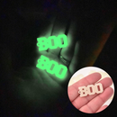 A hand in a dark room holding two glowing charms that say "BOO" in all caps. In the bottom right corner, a small circle shows a hand holding the same charm in a bright room; the green glow is now a pale cream.