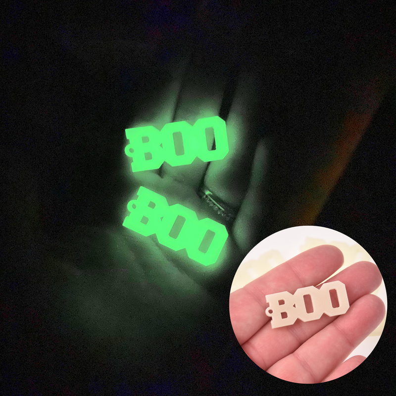 A hand in a dark room holding two glowing charms that say "BOO" in all caps. In the bottom right corner, a small circle shows a hand holding the same charm in a bright room; the green glow is now a pale cream.